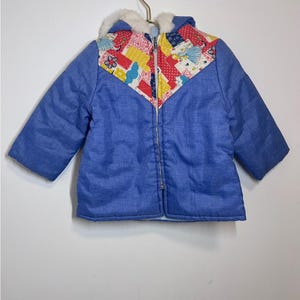 May include: A blue quilted jacket with a patchwork design on the front. The jacket has a hood lined with white faux fur and a zipper closure.