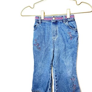 Könnte beinhalten: Hellblaue Jeanshose mit lila Blumenstickerei. Die Hose hat einen lila-weiß gemusterten Bund und hängt an einem goldfarbenen Kleiderbügel. Die Beinöffnungen sind ausgestellt.