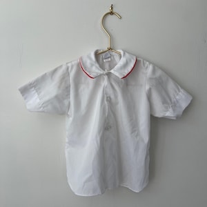 Puede incluir: Camisa blanca de manga corta con botones y ribete rojo alrededor del cuello.