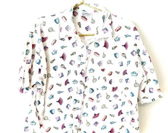 May include: White button-down shirt with a repeating pattern of small, colorful illustrations of fans, compacts, and other vintage items. The shirt is hanging on a gold hanger.