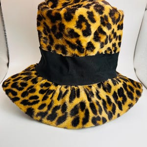 Op de afbeelding: Een hoed met luipaardprint en een zwarte band. De hoed heeft een imitatiebonttextuur met zwarte vlekken op een bruine achtergrond. De hoed heeft een brede rand en een cilindrische kroon.