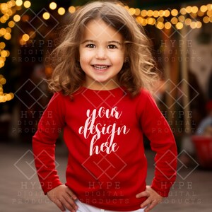 Puede incluir: Un niño pequeño con una camisa roja de manga larga con el texto "your design here" en blanco. El niño sonríe y está de pie frente a un fondo borroso de luces.