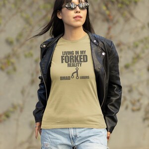 Puede incluir: Una camiseta color arena con el texto "LIVING IN MY FORKED REALITY" y "BMAD Losers" en negro. La camiseta se usa con una chaqueta de cuero negra y vaqueros azules.