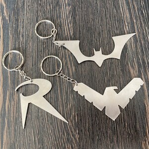 Peut inclure: Trois porte-clés en métal représentant les logos de Batman, Robin et Nightwing. Les porte-clés sont argentés et ont un anneau en haut pour attacher les clés.