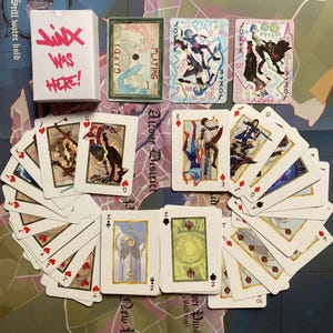 Peut inclure: Un jeu de cartes à jouer personnalisé avec des illustrations uniques. La boîte de cartes blanche porte l'inscription "Jinx Was Here!" en rose. Les cartes comportent les mots "Joker" et "Cards Playing". Les cartes sont étalées sur une carte.