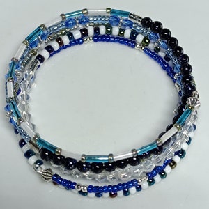May include: A multi-strand wrap bracelet featuring a mix of blue, white, and clear beads. The bracelet includes various bead sizes and shapes, with silver-toned metal accents. The design incorporates a range of blues, from light to dark, creating a layered effect.