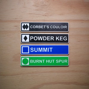 Può includere: Quattro cartelli rettangolari con sfondi di colori diversi e testo bianco. Il cartello superiore è nero con testo bianco che recita "Corbet's Couloir". Il secondo cartello è nero con testo bianco che recita "Powder Keg". Il terzo cartello è blu con testo bianco che recita "Summit". Il cartello inferiore è verde con testo bianco che recita "Burnt Hut Spur".