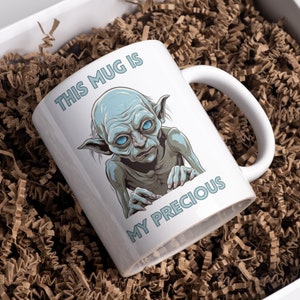 May include: White ceramic mug with a blue and green illustration of Gollum from the Lord of the Rings. The mug has the text "This mug is my precious" printed on it.