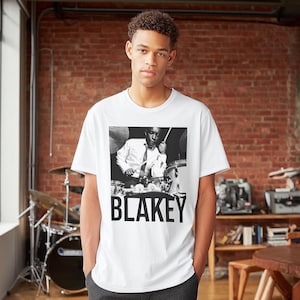 Puede incluir: Camiseta blanca con una imagen en blanco y negro de un baterista tocando la bater&iacute;a. La palabra "BLAKEY" est&aacute; impresa en letras negras en negrita debajo de la imagen. La camiseta la lleva una persona.