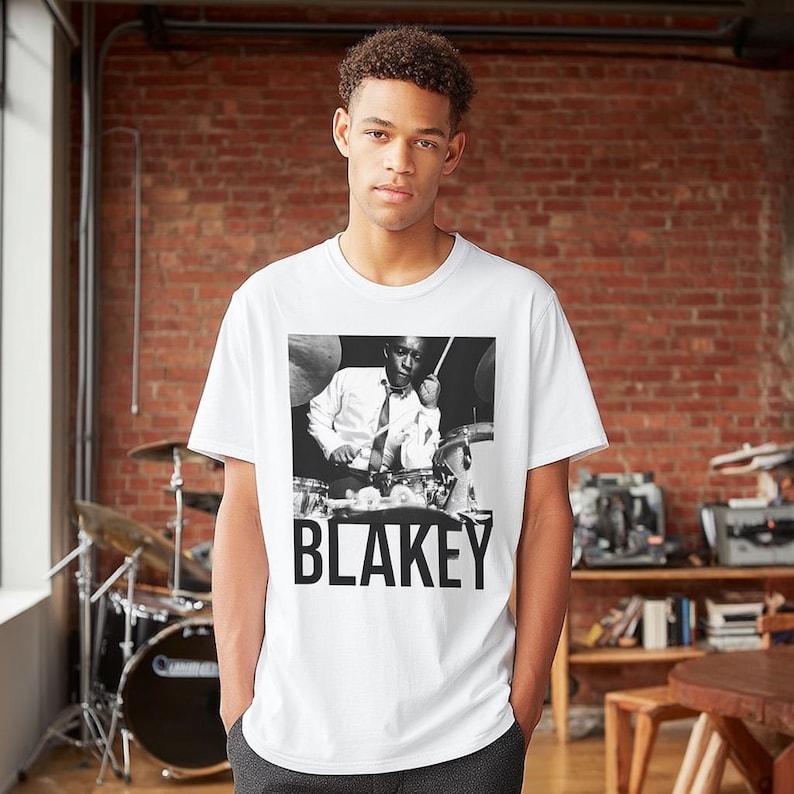 Puede incluir: Camiseta blanca con una imagen en blanco y negro de un baterista tocando la bater&iacute;a. La palabra "BLAKEY" est&aacute; impresa en letras negras en negrita debajo de la imagen. La camiseta la lleva una persona.