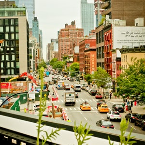 May include: A view of a city street with tall buildings and traffic. The street is lined with trees and there are cars parked on both sides of the street. The street is busy with people walking and driving.