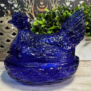 May include: A cobalt blue glass hen-on-nest dish. The hen is detailed with textured feathers and sits atop a woven basket-like base. This decorative piece is a charming addition to any kitchen or home decor.