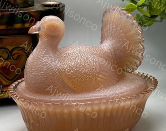 May include: A peach-colored glass bird figurine with a detailed tail, perched atop a matching dish. The bird has textured feathers and a delicate beak. The dish has a ribbed design, creating a vintage aesthetic.