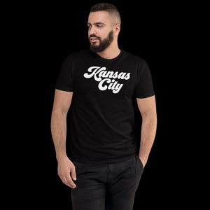 May include: Black t-shirt with the white text "Kansas City" in a retro script. The shirt is short-sleeved and appears to be made of a soft material. The person is wearing black trousers. The background is black.