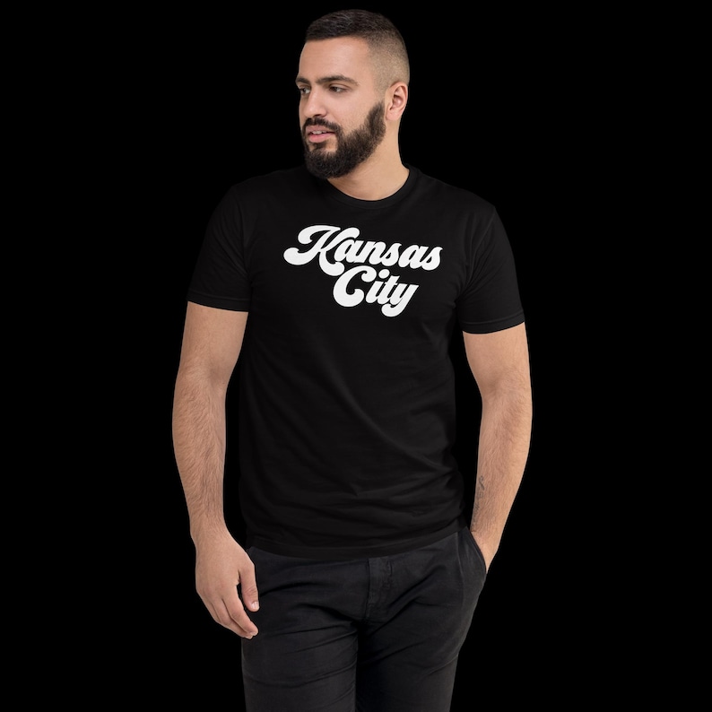 May include: Black t-shirt with the white text "Kansas City" in a retro script. The shirt is short-sleeved and appears to be made of a soft material. The person is wearing black trousers. The background is black.