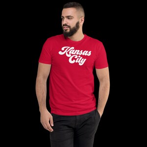 May include: A red t-shirt with the words "Kansas City" in white, stylized script. The shirt is short-sleeved and appears to be made of a soft material. The person is wearing black trousers.