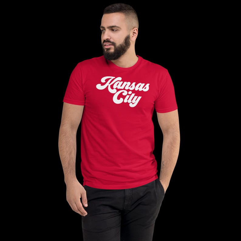 May include: A red t-shirt with the words "Kansas City" in white, stylized script. The shirt is short-sleeved and appears to be made of a soft material. The person is wearing black trousers.