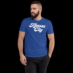 May include: Royal blue t-shirt with the words "Kansas City" in white, stylized script. The shirt has short sleeves and a crew neck. The person in the image is wearing black trousers.