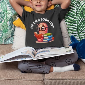 Op de afbeelding: Een jonge jongen draagt een grijs T-shirt met een cartoonworm die een boek leest en de tekst "I'm on a book diet". Hij zit op een bank met een boek op schoot.