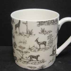 May include: White ceramic mug featuring a black and white pattern of African animals, such as a giraffe, zebra, elephant, and antelope. The mug has a large handle.