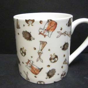 May include: White ceramic mug decorated with a playful design of cartoon woodland creatures. The illustrations feature foxes, hedgehogs, owls, and rabbits in brown, orange, and beige. The mug has a white handle and a shiny surface.
