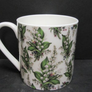 May include: White ceramic mug with a floral pattern of lily of the valley flowers and leaves. The mug has a white handle.