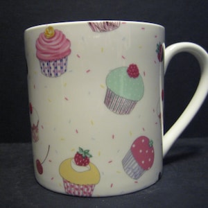 May include: White ceramic mug with a handle, featuring a colourful cupcake design. The mug is decorated with various cupcakes, sprinkles and cherries.