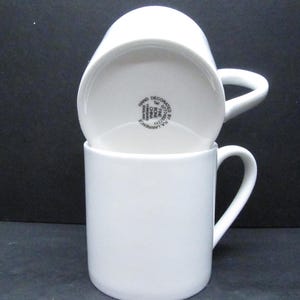 May include: Two white ceramic mugs, one stacked on top of the other. The top mug is upside down, displaying the base with the text "HAND DECORATED BY R.A. LAWRENCE". Both mugs have a curved handle. The mugs are made of fine bone china.