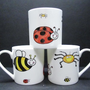 May include: Three white ceramic mugs with cartoon insect designs. One mug features a ladybug, another a bee, and the third a spider. Each mug has a white handle and a black background.