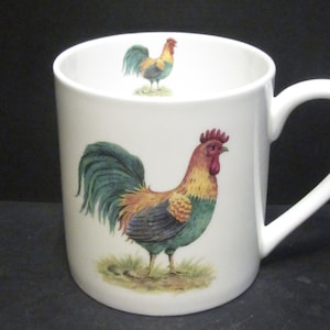 May include: White ceramic mug featuring a rooster illustration. The rooster is depicted in shades of green, red, yellow, and black. The mug has a white handle.