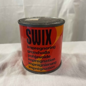 Può includere: Una lattina rossa e arancione con la parola "SWIX" in lettere nere. La lattina ha testo in più lingue, tra cui "impregnering", "grundvalla", "pohjavoi", "impregnation", "imprägnierung" e "imprégnation".