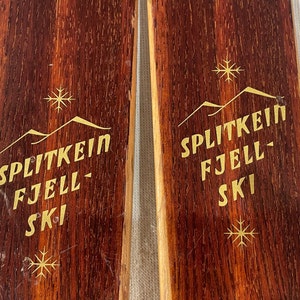 Puede incluir: Un par de esquís de madera vintage con letras doradas que dicen "Splitkein Fjell Ski" y una cordillera estilizada con copos de nieve sobre el texto.