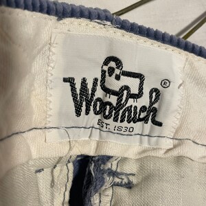 May include: A white label with black text and a cartoon sheep logo. The label says "Woolrich EST. 1830". The label is attached to a pair of blue corduroy pants.