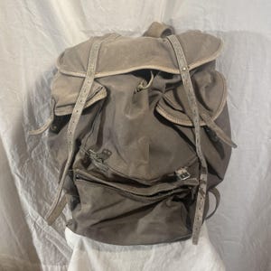May include: A large, gray canvas backpack with leather straps and buckles. The backpack has a flap closure and multiple pockets.