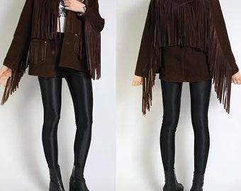May include: A brown suede jacket with long fringe detailing on the sleeves and back. The jacket has a button-up front and two front pockets. The model is wearing black leggings and platform boots. The jacket is a vintage style.