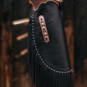 May include: Black leather chaps with a brown leather waistband and decorative silver conchos. The chaps feature long fringe along the sides and are accented with silver studs. A classic Western wear item.