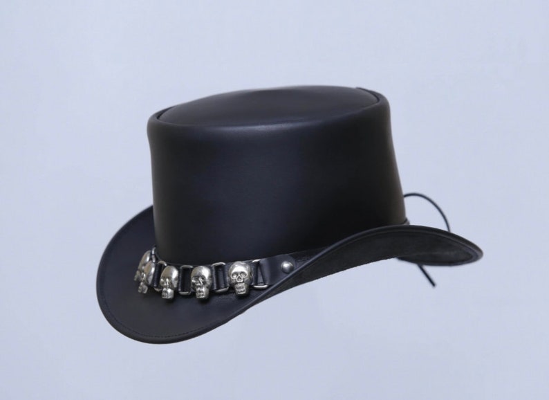 May include: A black leather top hat with a wide brim. The hat features a silver-colored band adorned with skull-shaped embellishments. The hat is a classic style, suitable for various occasions.