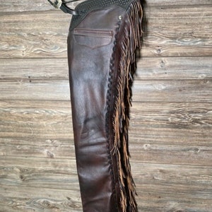 May include: Brown leather chaps with fringe detailing. The chaps have a black waistband with a decorative silver buckle and a pocket on the front. The fringe is a lighter brown colour.
