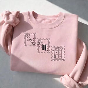 May include: A light pink crewneck sweatshirt featuring three black embroidered postage stamp designs. The left stamp shows butterflies, the center stamp displays the BTS logo, and the right stamp depicts a window with a plant and stars.