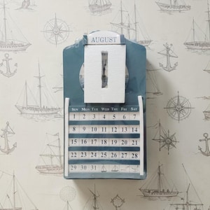 May include: A blue wall-mounted perpetual calendar featuring a clock face at the top with the month AUGUST printed above it. The calendar grid displays the days of the week and numbers 1 through 31, with small sailboat icons on the final row.
