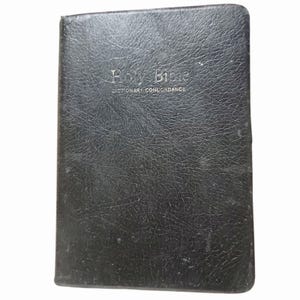 May include: A black, leather-bound Holy Bible. The cover is embossed with the words "Holy Bible" and "Dictionary Concordance" in gold. The leather has a textured finish and shows signs of wear.