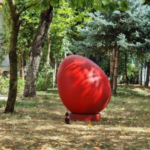 Könnte beinhalten: Eine große, rote, eiförmige Skulptur steht auf einem grasbewachsenen Rasen mit Bäumen im Hintergrund.