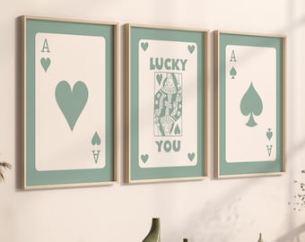 May include: Three framed prints with a green background and white card designs. The center print features a queen of hearts card with the text "LUCKY YOU". The left print features an ace of hearts card with a heart shape. The right print features an ace of spades card with a spade shape.