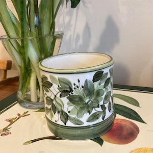 May include: A cylindrical ceramic planter with a white body and green trim. It features a hand-painted design of green flowers and leaves. A vase of flowers is visible in the background.