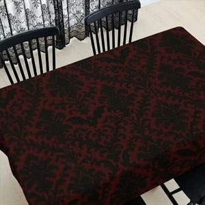 May include: A burgundy and black damask tablecloth covers a table, with two black chairs visible. The tablecloth features a repeating floral pattern in black against a burgundy background. The scene is set indoors, with a lace curtain in the background.