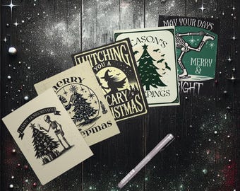 Puede incluir: Una colección de tarjetas navideñas espeluznantes con varios diseños, incluyendo un esqueleto decorando un árbol de Navidad, una bruja y un esqueleto patinando sobre hielo. Las tarjetas presentan frases como "Happy Horrordays" y "Merry & Bright". Un bolígrafo plateado descansa sobre la superficie oscura y moteada.
