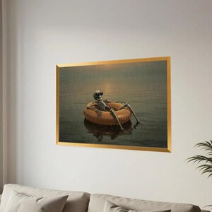 Op de afbeelding: Een ingelijste print toont een skelet met een zonnebril, ontspannen in een oranje opblaasbare ring in het water. De achtergrond toont een zonsondergang. De lijst is goudkleurig.