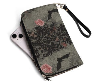 Puede incluir: Cartera gris con un patrón de damasco negro, rosas rosas y siluetas de murciélagos. La cartera tiene una correa de muñeca negra y una cremallera plateada. Un smartphone blanco es parcialmente visible en el interior.