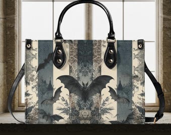 Puede incluir: Bolso tote rectangular con diseño de murciélago. El bolso tiene un asa y una correa para el hombro negras. La parte delantera del bolso presenta un patrón de rayas con ilustraciones de murciélagos y detalles florales en tonos grises, crema y negro.