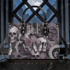 May include: A black handbag with a gothic design featuring skulls, roses, and butterflies in shades of gray and purple. The bag has a black handle and a detachable shoulder strap.